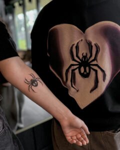 Tatuaż oldschool przedstawiający małego pająka na kobiecym przedramieniu. Styl tatuatorski z wyraźnymi konturami. Tatuaż umieszczony w widocznym miejscu na ciele, idealny przykład damskiego tatuażu ze zwierzęcym motywem.